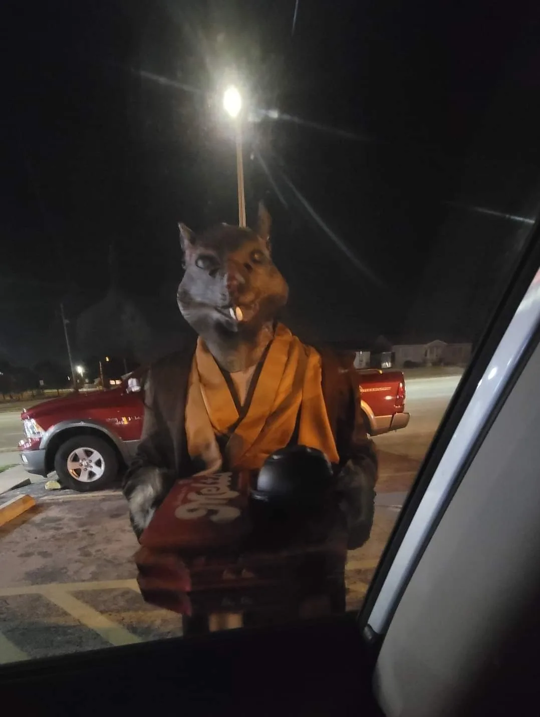 A person wearing a large, realistic rat mask and a brown robe with an orange sash stands outside at night, holding a tray…