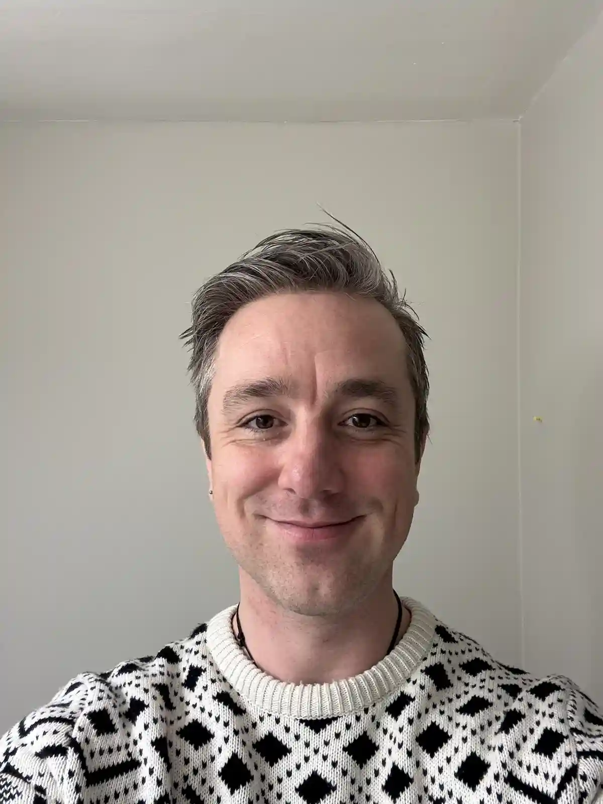 A man with salt-and-pepper hair and a subtle smile wears a black-and-white patterned sweater against a plain wall. The…