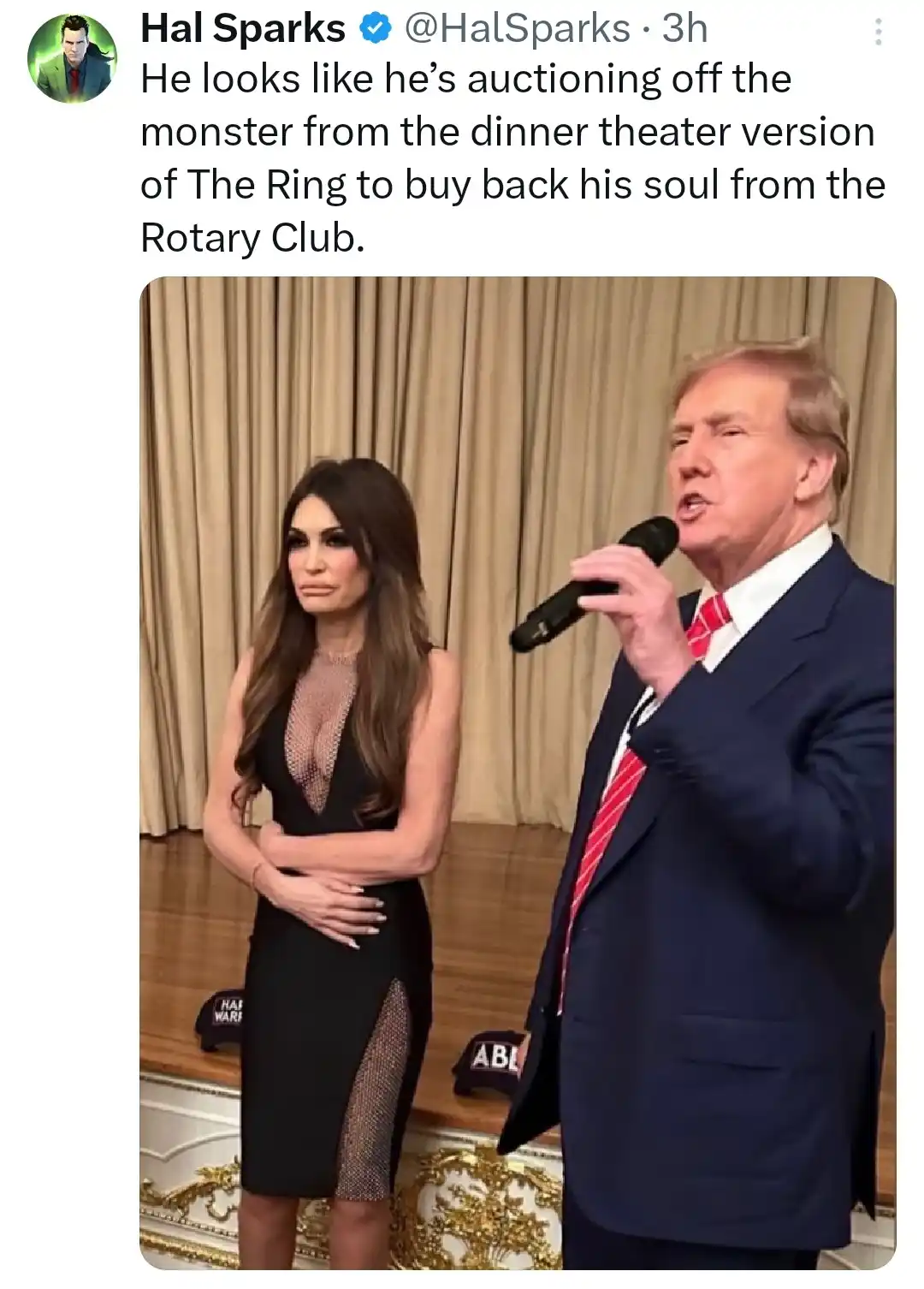 A man in a dark suit holding a microphone speaks while a woman in a black dress stands beside him with her arms crossed. The…