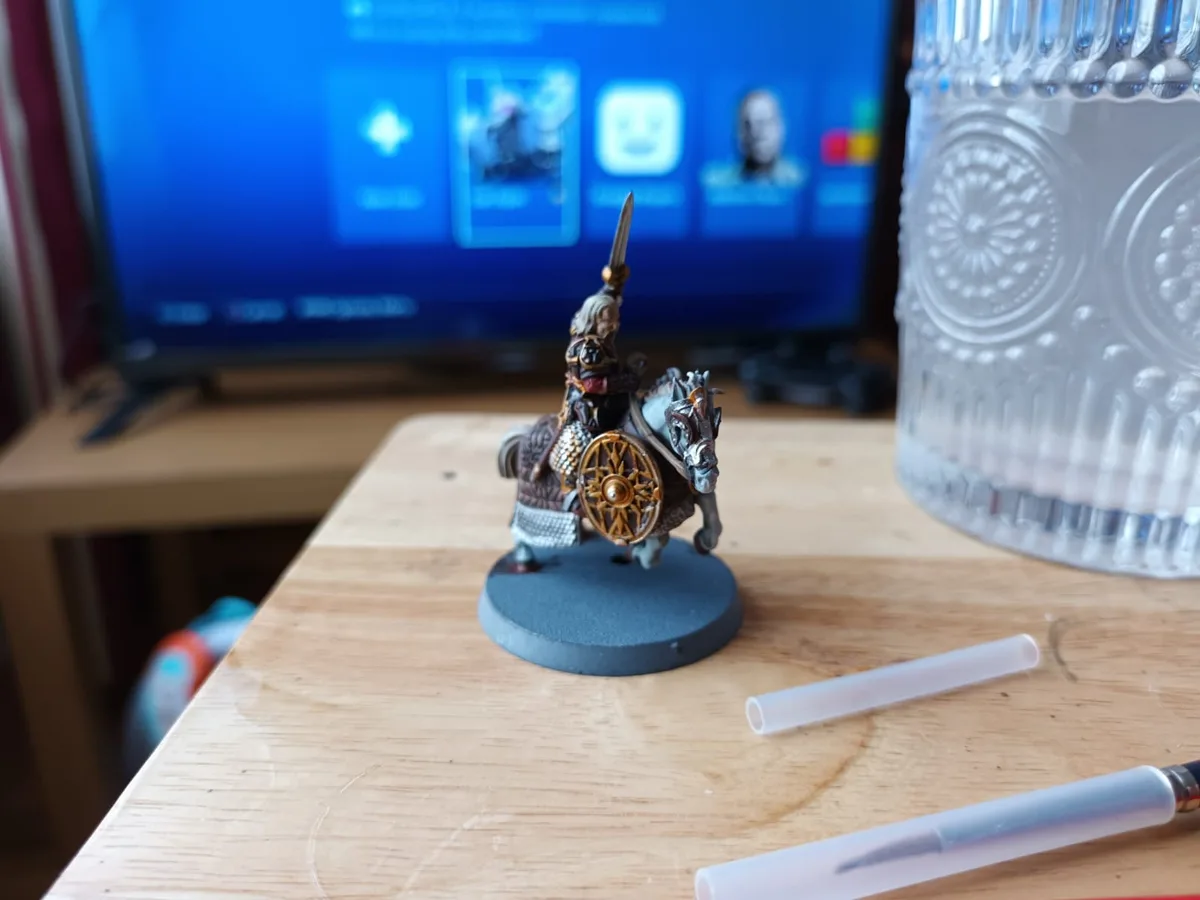 A painted miniature of a mounted warrior holding a sword and shield stands on a wooden table next to paintbrushes and a…