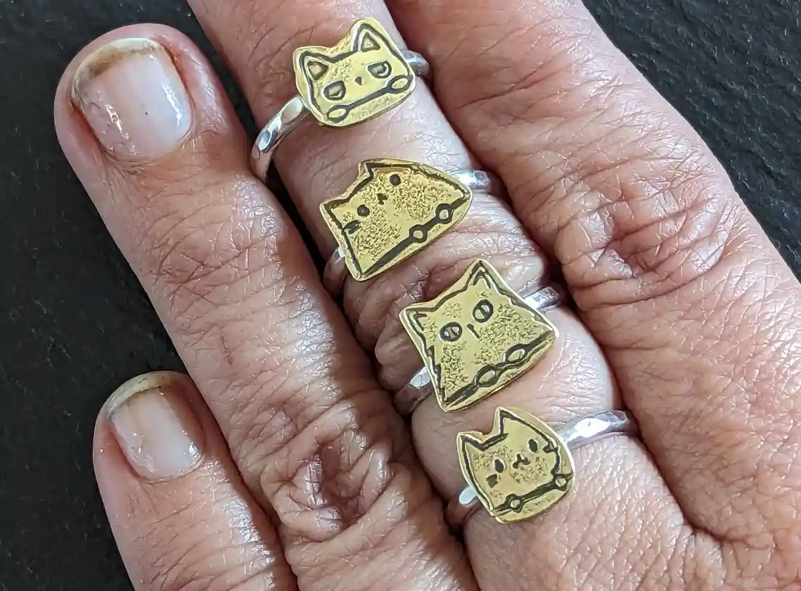A close-up photo shows a person's hand with four rings, each featuring a small, stylized gold cat face design, worn on…
