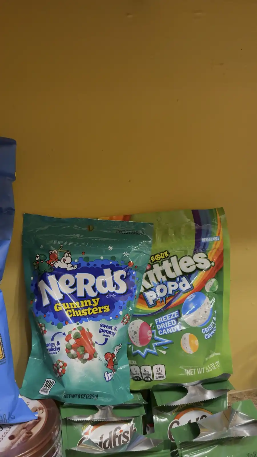 Two bags of candy, Nerds Gummy Clusters (green bag) and Sour Skittles POP'd (green bag), sit side by side against a yellow…