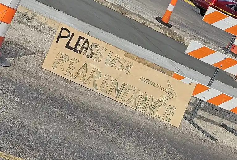 A handmade wooden sign on a road with orange and white traffic barriers and cones, reading 'PLEASE USE REAR ENTRANCE' with…