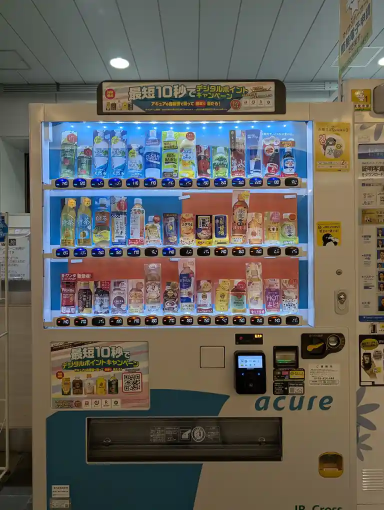 A brightly lit Japanese vending machine branded 'acure' displays multiple rows of beverages with price tags, featuring…