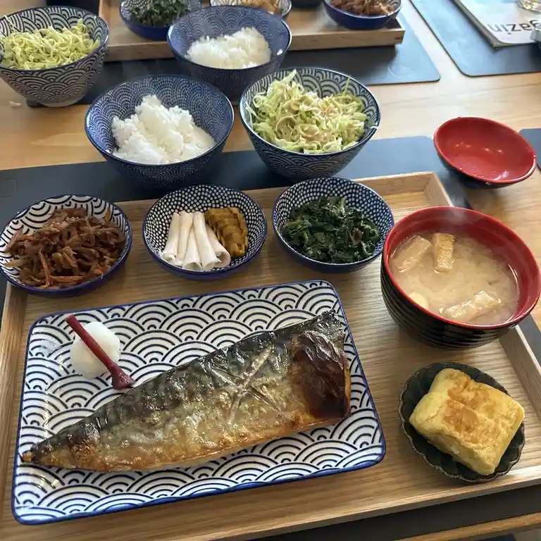 A Japanese meal featuring grilled fish, rice, miso soup, pickles, and side dishes arranged on a wooden tray. The overlaid…