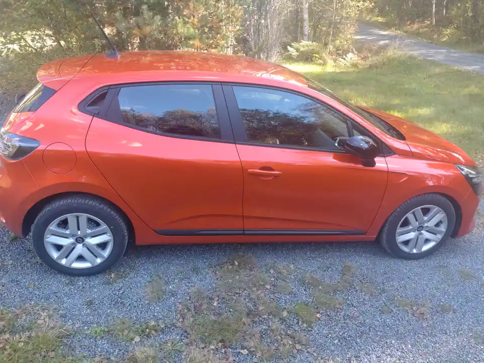 A bright orange Renault Clio hatchback is parked on a gravel driveway with grass and trees in the background. The car is…