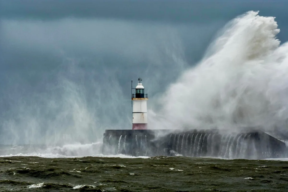 A lighthouse stands resiliently on a pier as an enormous, crashing wave explodes against it, sending water spraying high…