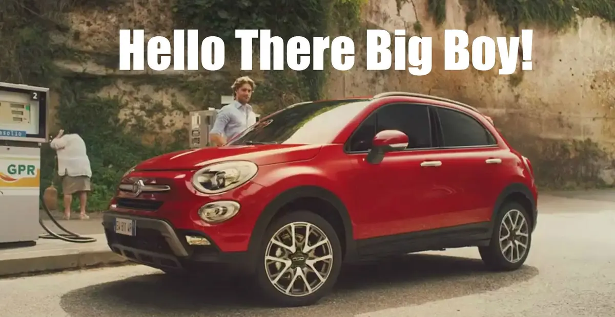 A red Fiat 500X SUV is parked at a gas station with a man in a light blue shirt standing beside it. In the background…