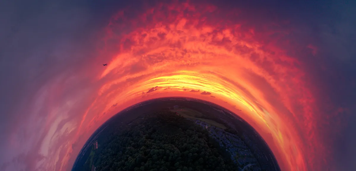 A fisheye lens photo shows a dramatic sunset with fiery red and orange clouds arching over a dark landscape of trees and…