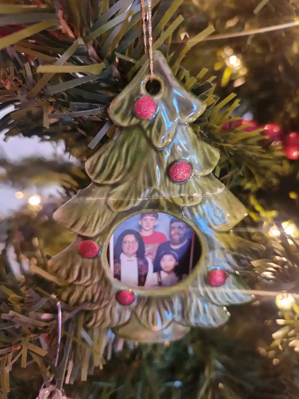 A ceramic Christmas tree ornament with red glitter dots hangs on a tinsel tree, featuring a family photo inside its central…