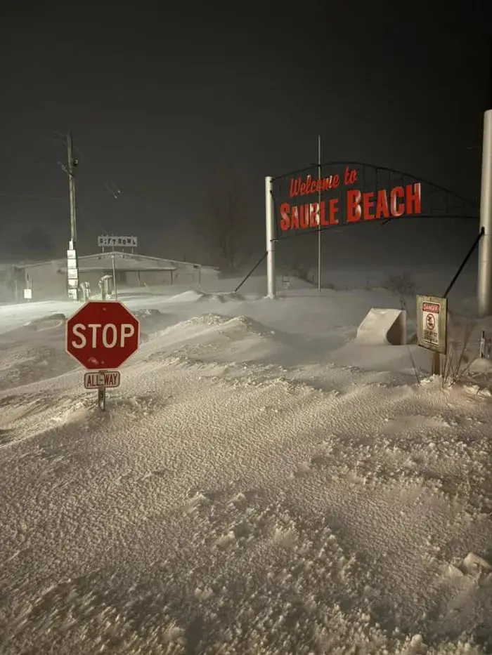 A nighttime photo shows a snow-covered street with a red stop sign and an 'ALL-WAY' plaque partially buried in snow. In the…