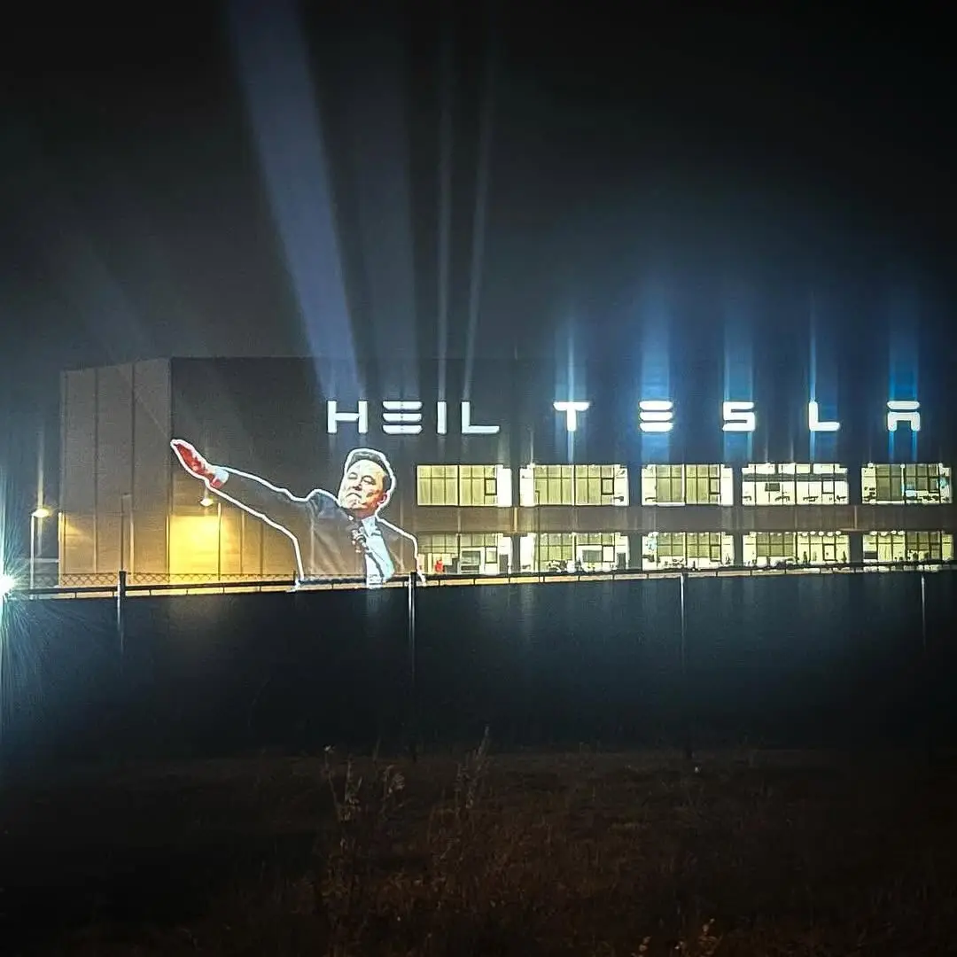 A nighttime photo of a large building with illuminated signage reading 'HEIL TESLA'. Projected onto the side of the building…