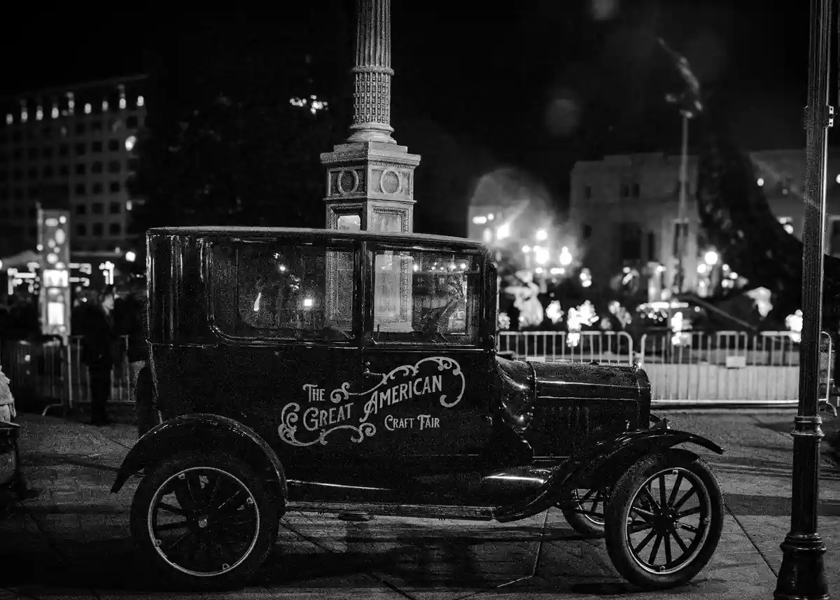 A black-and-white photo of a vintage 1920s-style automobile parked at night, with ornate lettering on its side reading 'The…