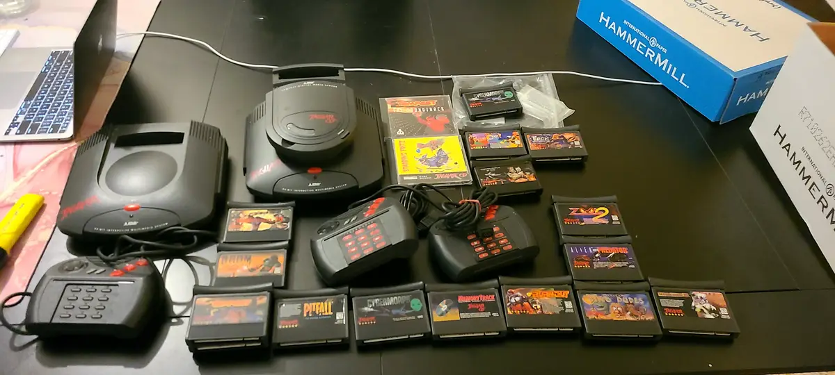 A black Atari Jaguar gaming console and its accessories are displayed on a dark table, surrounded by numerous game…