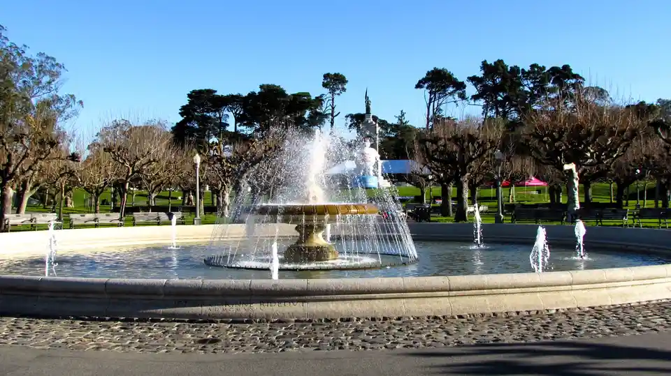 A wide shot of a large, ornate public fountain in a park with water jets spraying upwards and outwards. Behind the fountain…