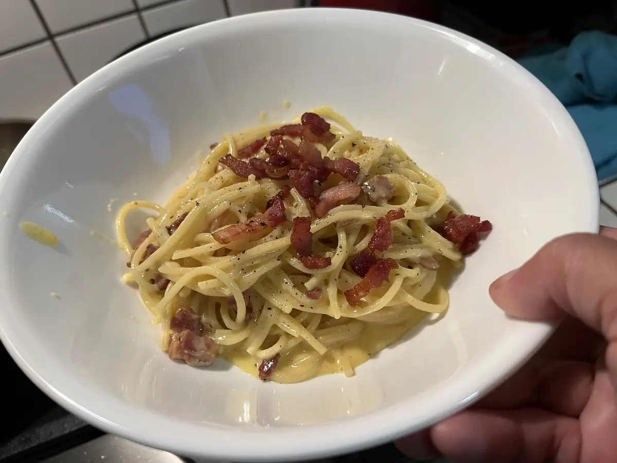 A hand holds a white bowl filled with creamy pasta generously topped with crispy bacon bits and black pepper, set against a…