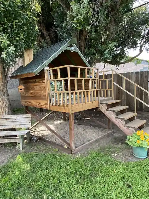 A wooden treehouse with a green roof and stairs, built into the branches of a large tree in a backyard with grass, a bench…