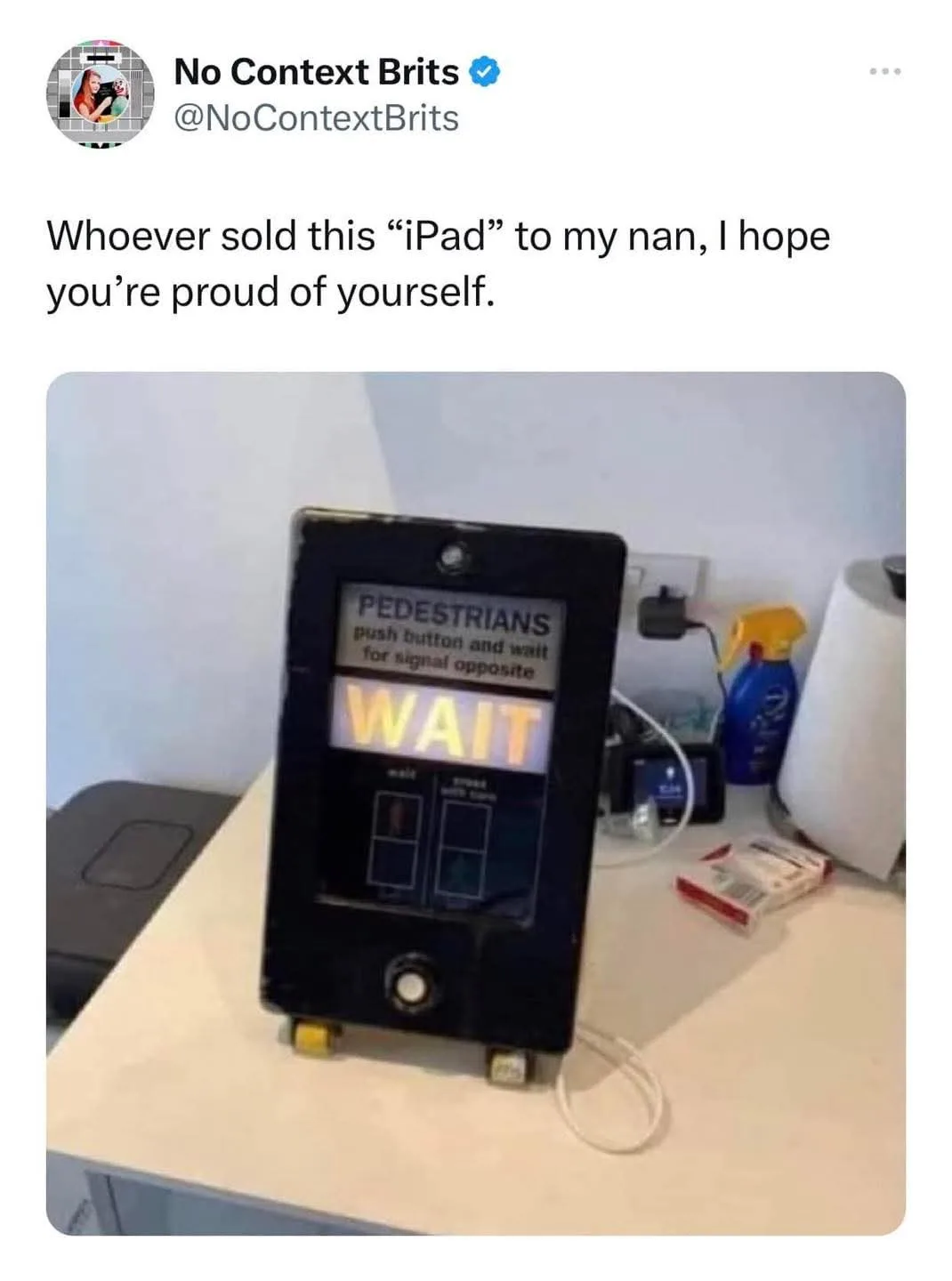A black, rectangular pedestrian crossing signal, resembling an iPad, sits on a countertop. Its screen displays 'PEDESTRIANS…