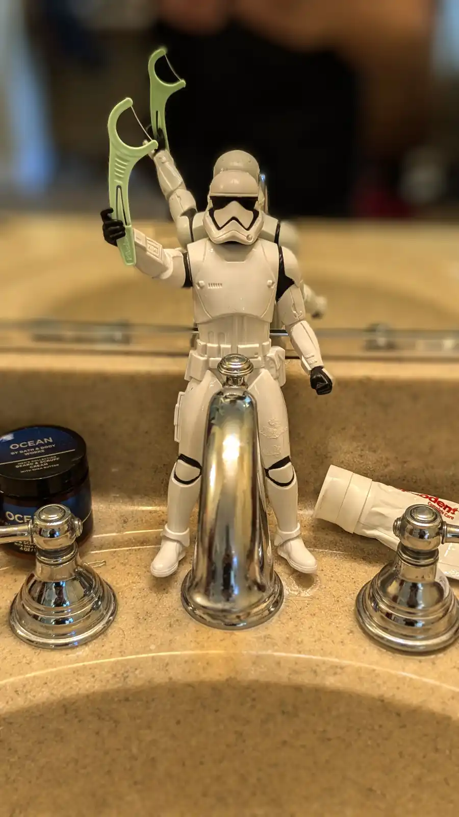 A white Star Wars Stormtrooper action figure stands on a bathroom sink counter, holding a green floss pick in both hands as…