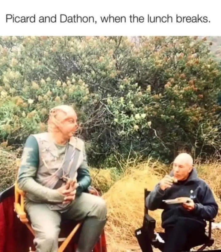 Two men sit in director's chairs outdoors, one in costume as the alien Dathon with a bald head and pointed ears, the other…