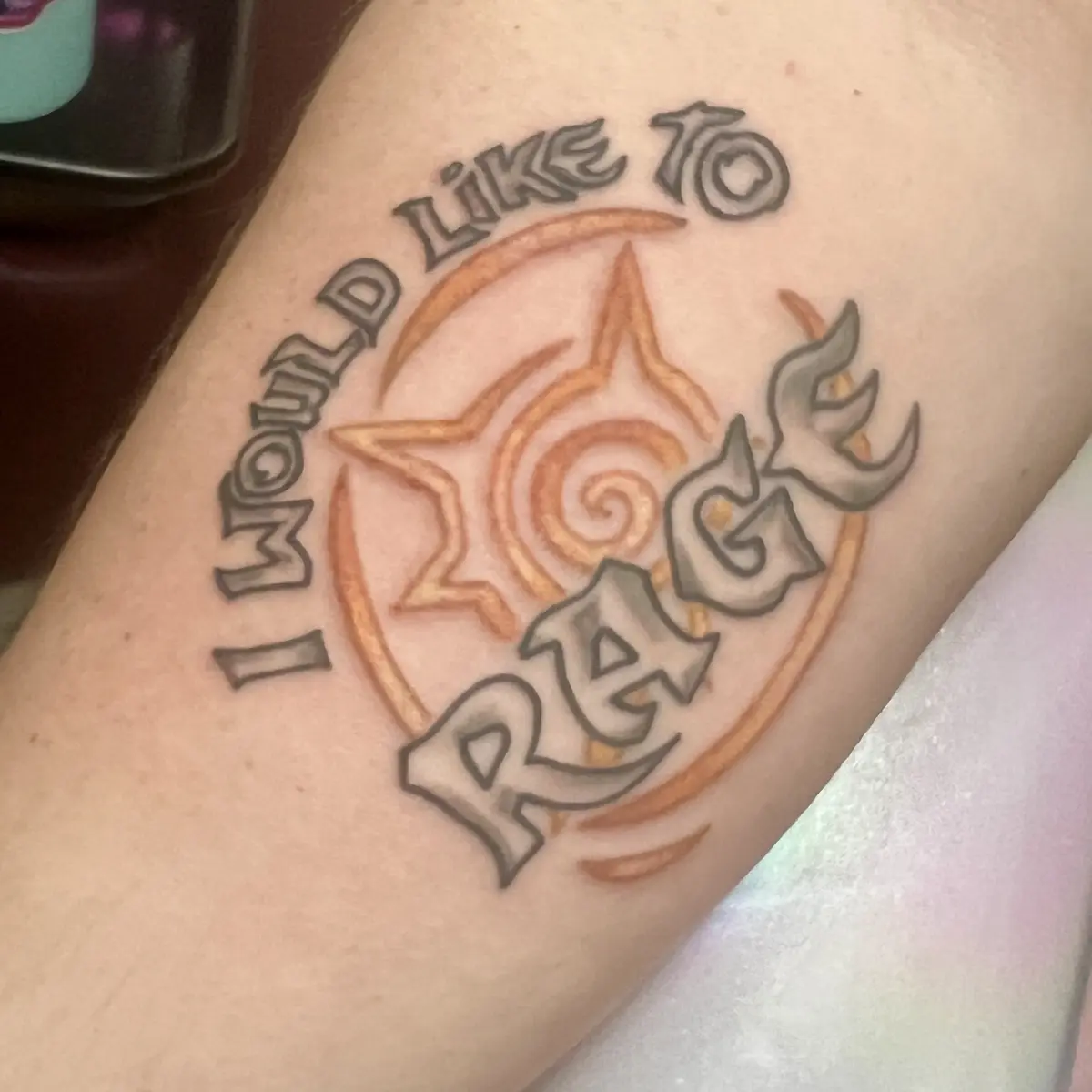 A close-up photo shows a tattoo on human skin, likely an arm, featuring the text 'I WOULD LIKE TO RAGE' in stylized…