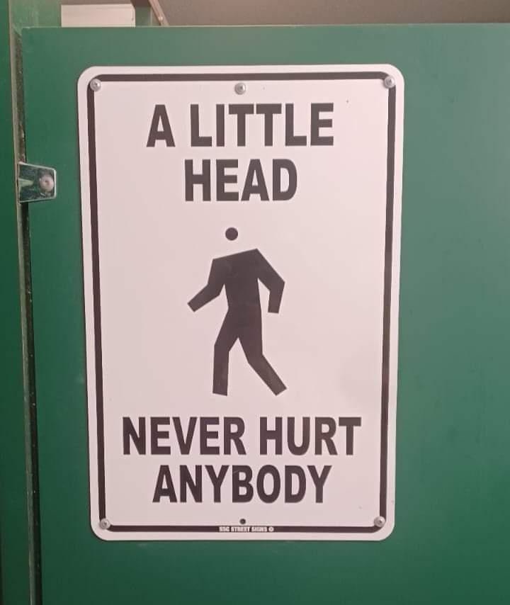 A white rectangular sign with a black border is mounted on a green door. The sign features the text 'A LITTLE HEAD' at the…