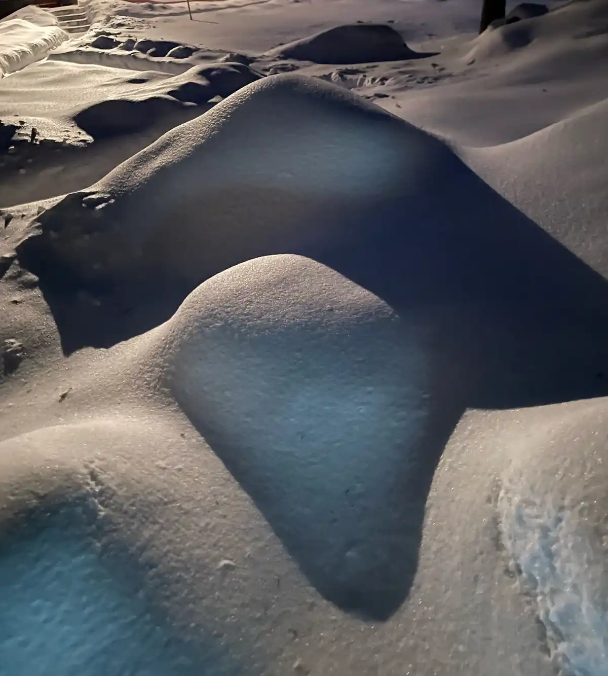 A close-up photo of snowdrifts with smooth, rounded mounds and deep shadows cast by low-angle sunlight. The snow's texture…