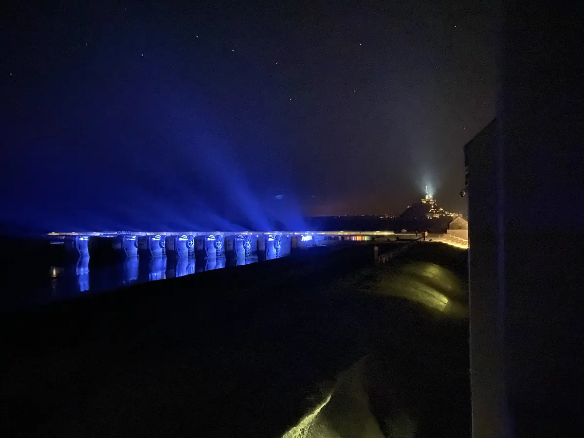 A nighttime photograph shows a long pier or breakwater illuminated by bright blue spotlights projecting beams into the dark…