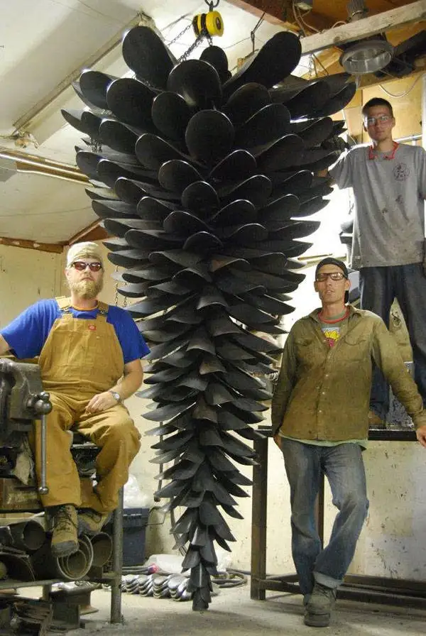 Three men stand in a workshop beside a giant sculpture made of stacked boat propellers, shaped like a pinecone. One man sits…