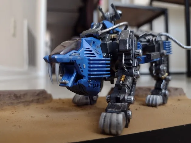 A blue and grey mechanical lion-like robot model with articulated legs and claws, standing on a sandy surface. The overlaid…