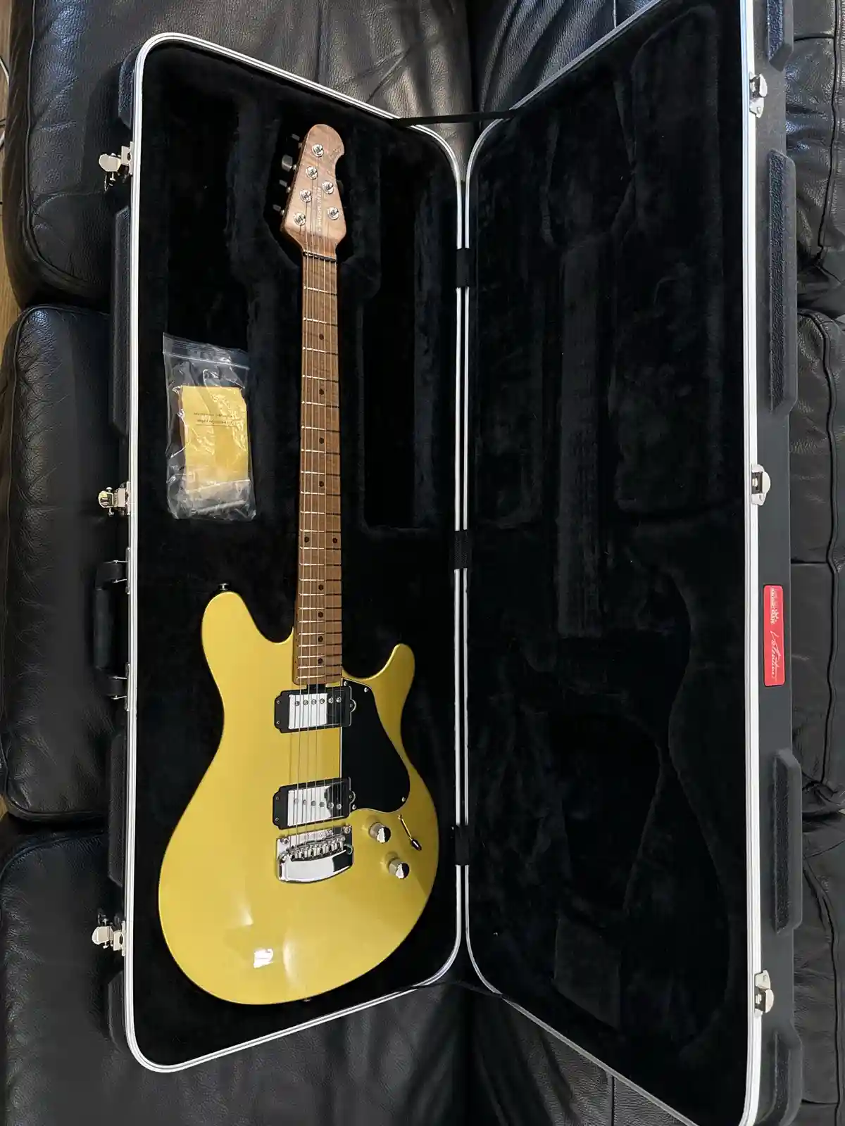 A yellow electric guitar sits inside an open black hardshell case resting on a black leather couch. A small plastic bag with…