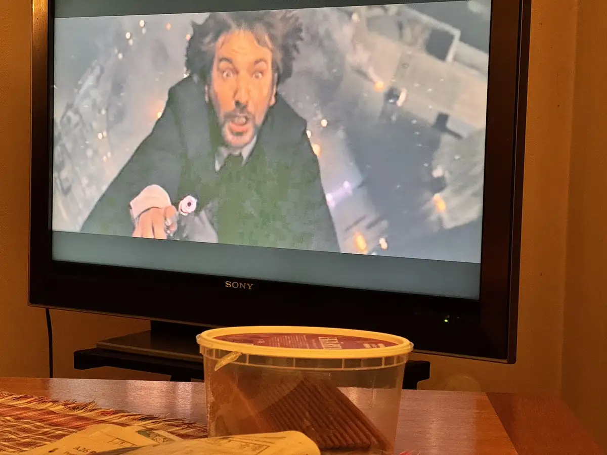 A television screen displays a close-up of a man with wild hair and an exaggerated shocked expression, wearing a dark coat…