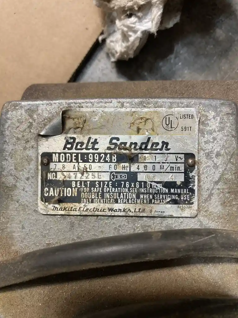 A close-up of a weathered, dusty metal label on a Makita belt sander, showing model number 9924B, voltage, amperage, belt…