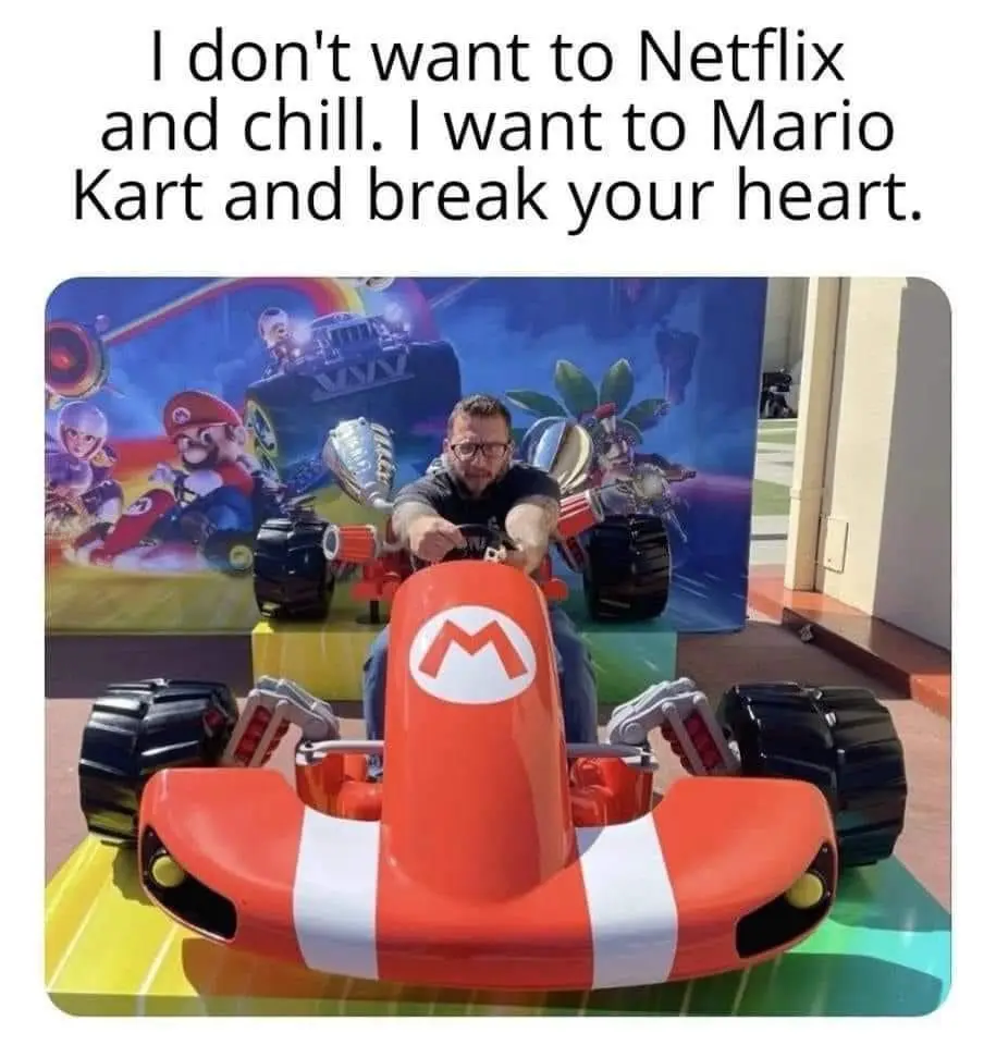 A man with glasses sits in a red Mario Kart ride-on toy car, gripping the steering wheel. Behind him is a colorful Mario…