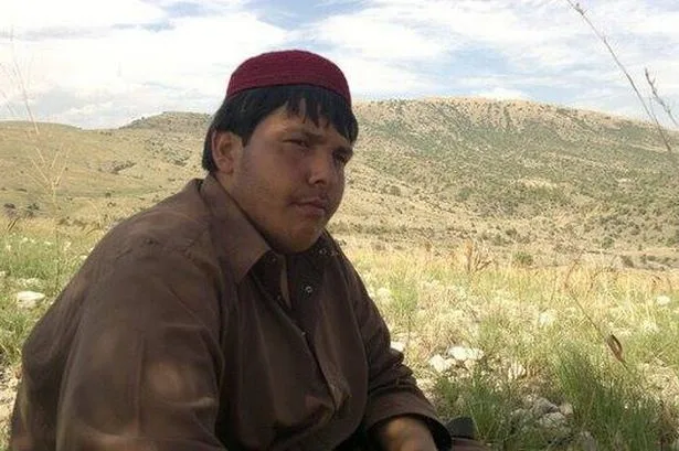 a man wearing a red cap and brown shirt sits in a grassy field with hills in the background, looking directly at the camera…