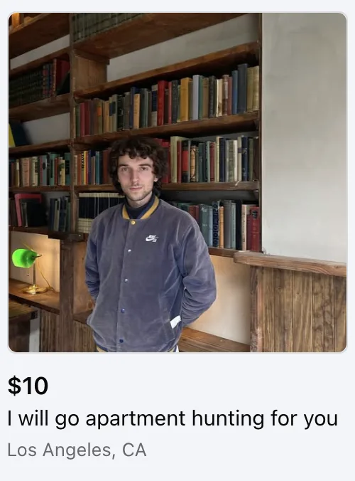 A young man with curly hair stands in front of a large wooden bookshelf filled with books. He wears a grey corduroy jacket…
