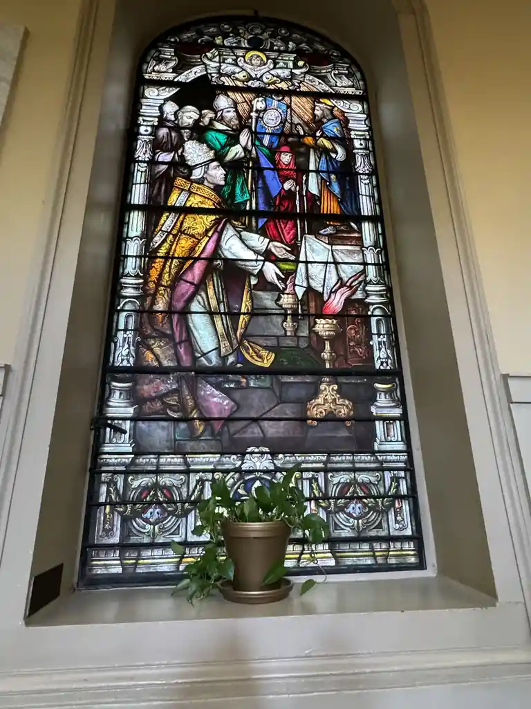 A stained glass window depicting a religious scene with figures in ornate robes, set within an arched frame. In front of the…