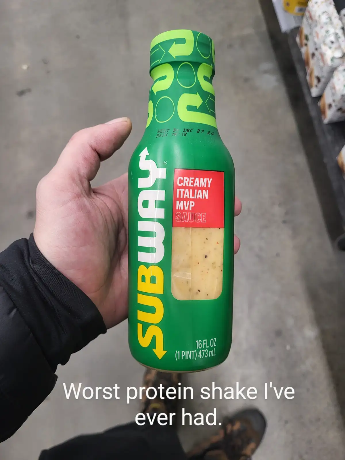 A hand holds a green Subway bottle labeled 'CREAMY ITALIAN MVP SAUCE' in a store aisle. The bottle has a visible 'BEST BY'…