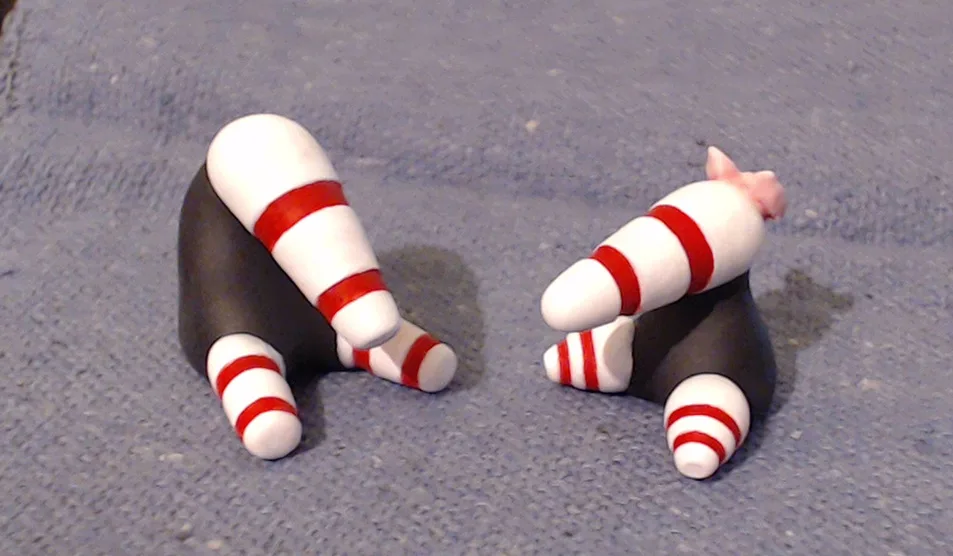 Two handmade clay figurines resembling candy cane-striped legs and feet, each with a black torso, are positioned on a gray…