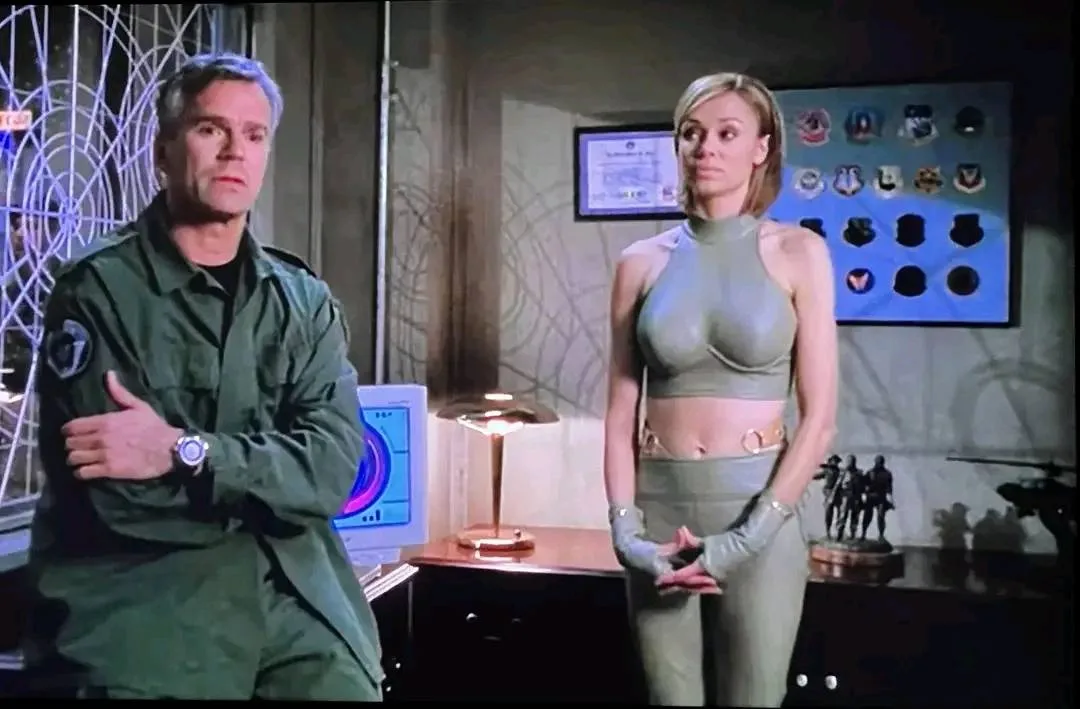 A man in a green military-style uniform stands with arms crossed, looking sternly at the camera. Beside him, a woman in a…