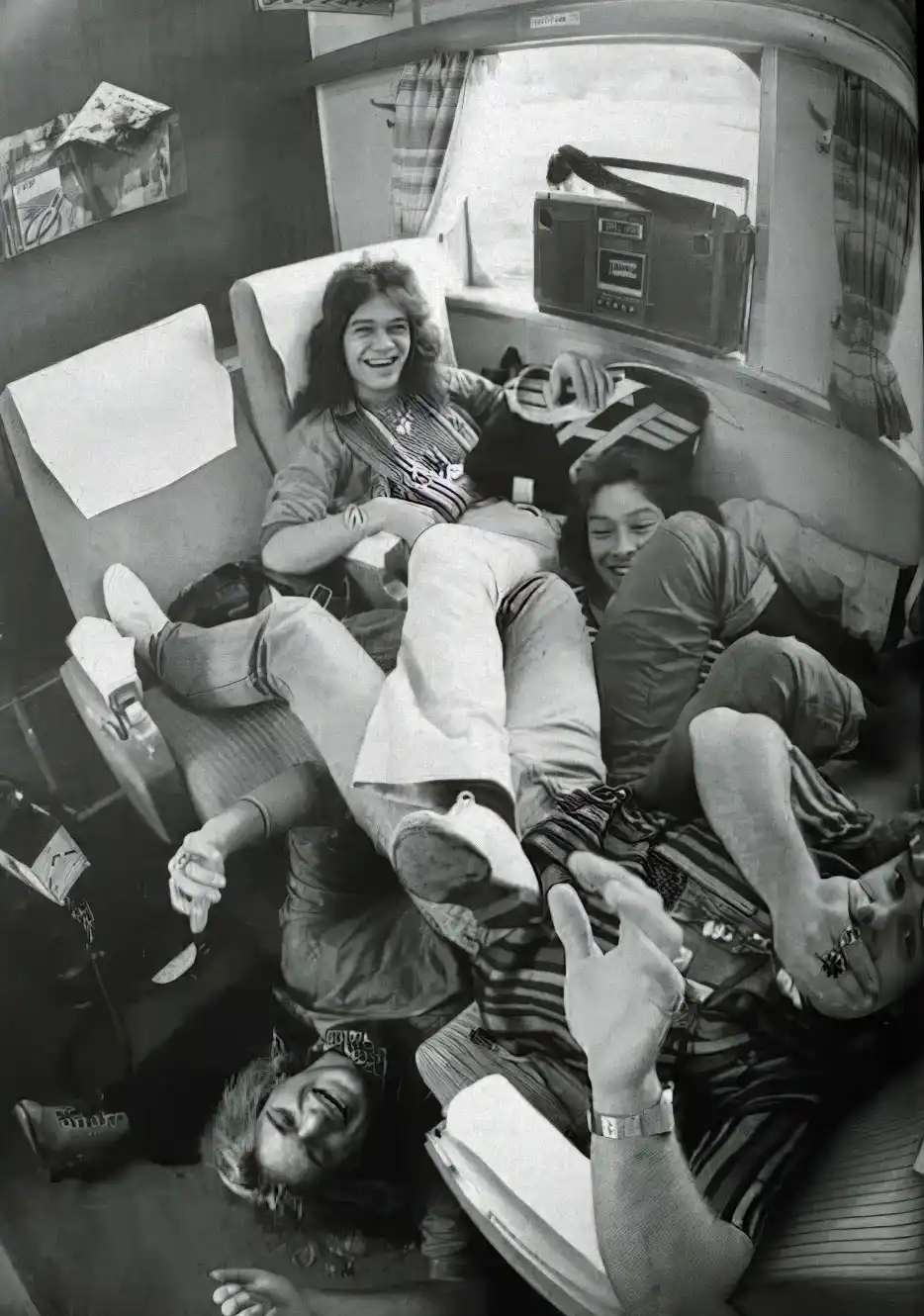 A black-and-white photo shows four young people lounging and laughing inside a vintage camper van, with one person making a…