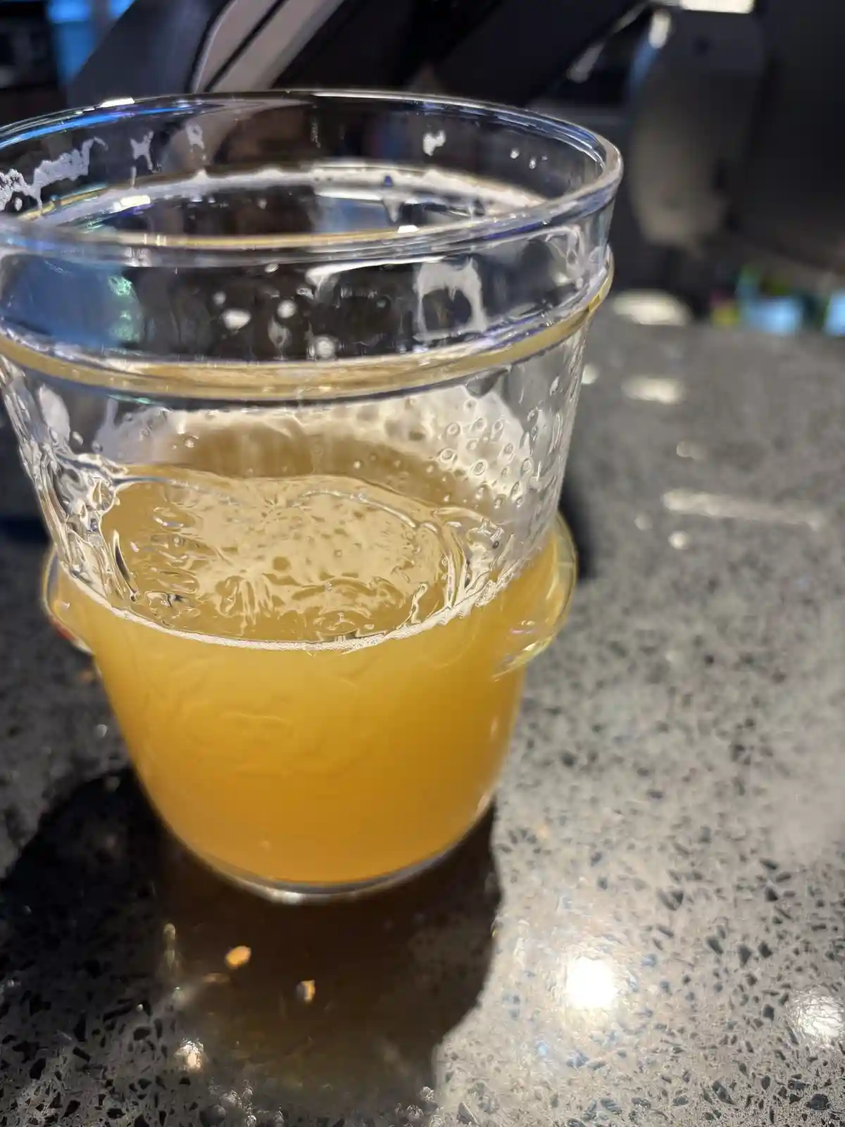 A close-up photo shows a clear glass filled with a golden-yellow beverage, likely beer, sitting on a dark speckled…