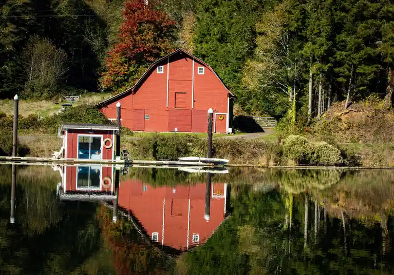 A serene landscape photo shows a red barn and a small boathouse on the edge of a calm body of water, with their reflections…