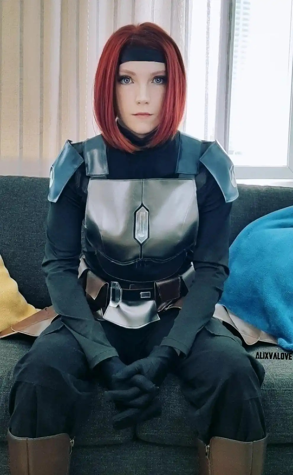 A person in a detailed cosplay of a female character with short red hair, black headband, and dark armor sits on a couch…