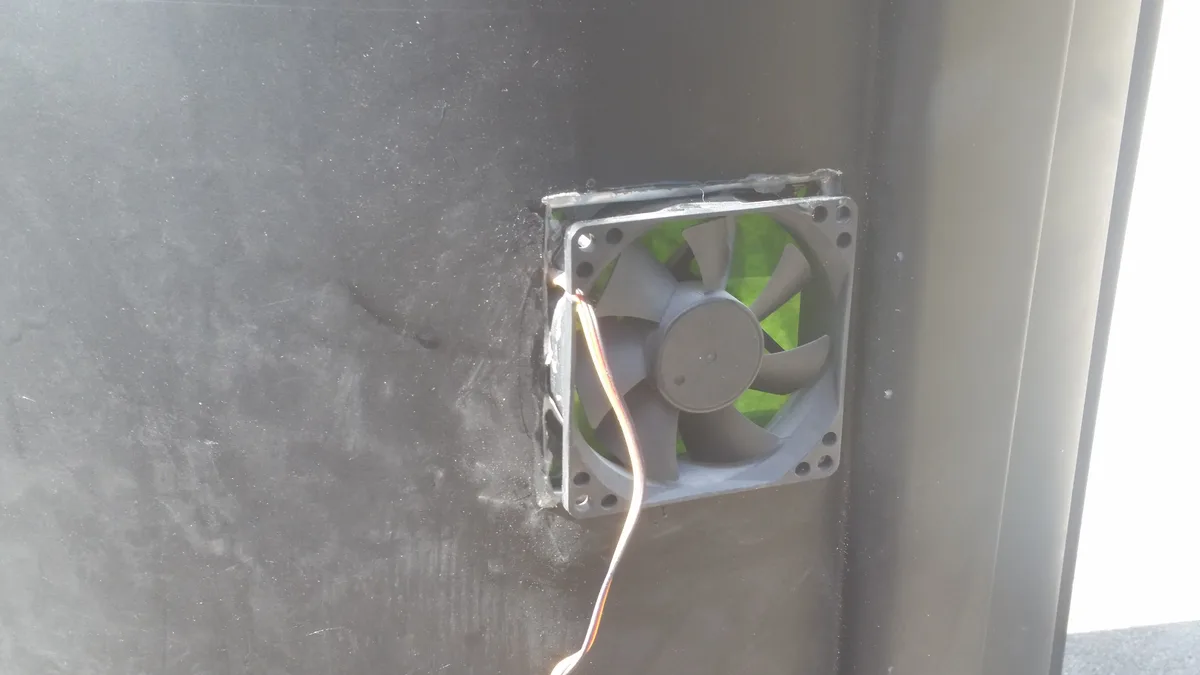 A square computer cooling fan is crudely mounted to a dark, dusty metal surface with visible wires extending from it…