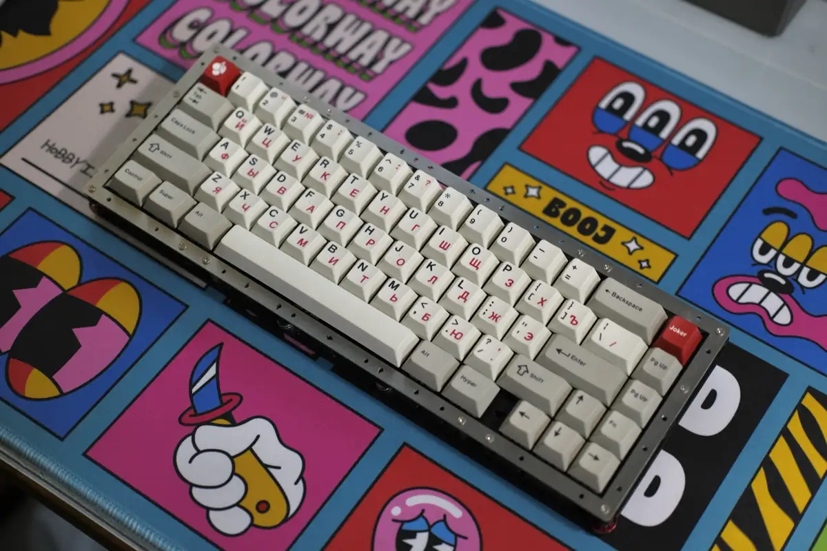 A mechanical keyboard with white and gray keys featuring Cyrillic and Latin characters sits on a colorful, cartoon-themed…