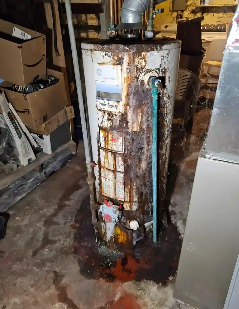 A severely rusted and leaking water heater stands in a cluttered basement, with brown water pooling on the concrete floor…