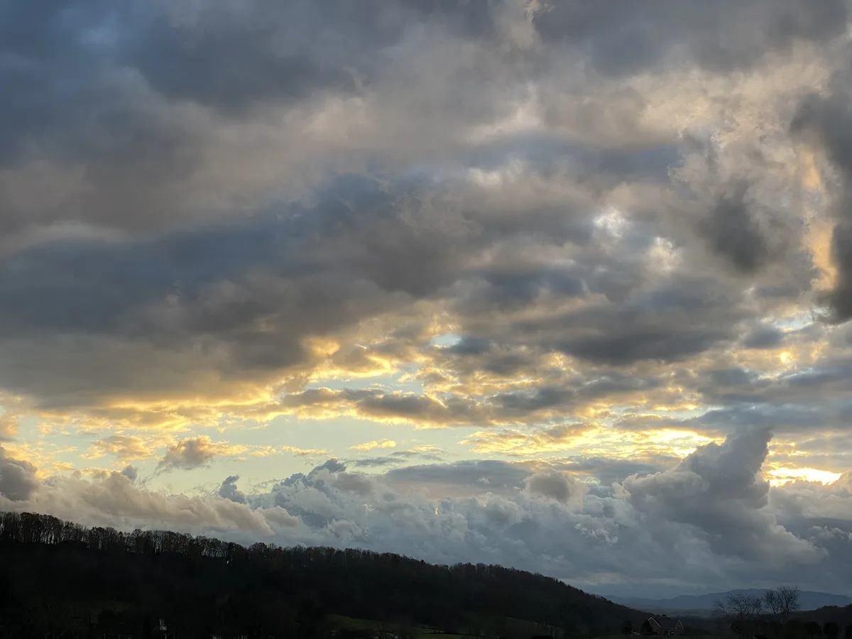 A dramatic landscape photo shows a cloudy sky at sunset or sunrise, with golden light breaking through the clouds and…