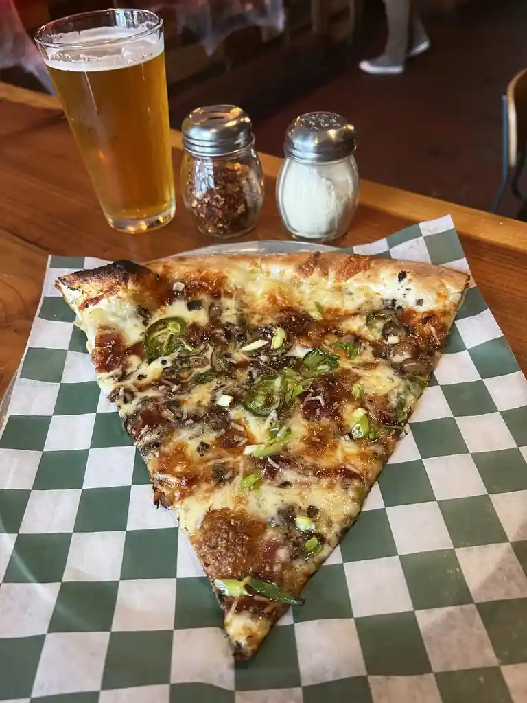 A single slice of pizza with melted cheese, green onions, and mushrooms sits on green-and-white checkered paper on a wooden…