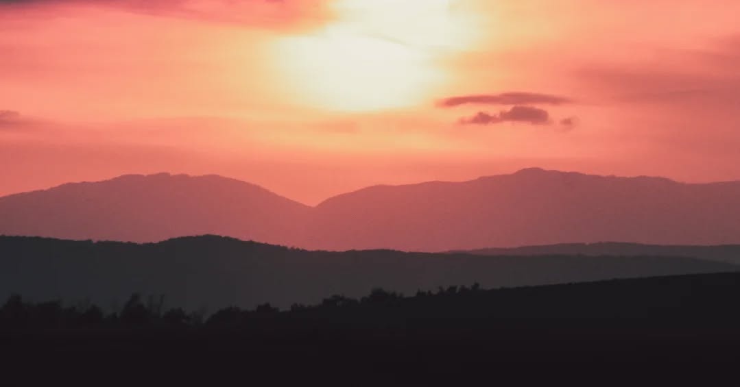 A serene landscape photo of a sunset over layered mountain ranges, with the sky glowing in warm orange and pink hues. The…